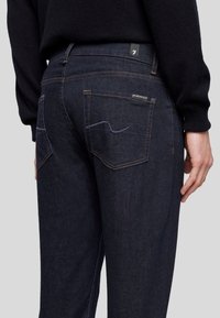 Donkerblauwe denim jeans met een getailleerd ontwerp, voorzien van achterzakken en subtiele paarse stiksels. Label zichtbaar bij de tailleband.