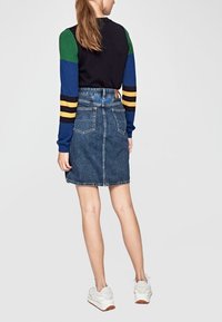 Kvinna klädd i en färgblockerad långärmad tröja, högmidjade denimkjol och vita sneakers, stående med ryggen vänd mot betraktaren.