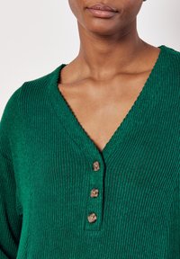 Top a coste verde con scollo a V, caratterizzato da tre bottoni marroni frontali e un tessuto testurizzato.