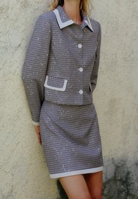 Szary i biały garnitur w kratę z kołnierzową, skróconą marynarką z guzikami oraz dopasowaną spódnicą, obydwa elementy z białymi akcentami wykończenia.