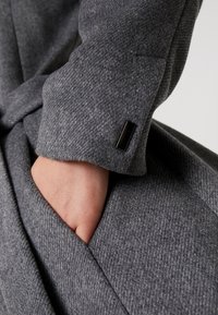 Cappotto in misto lana grigio con un tessuto texture, un elegante dettaglio in metallo nero sul polsino e una tasca laterale, con una vestibilità rilassata.