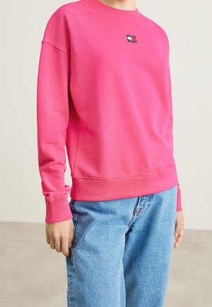 Rosa sweatshirt med långa ärmar och ribbad mudd, med en liten logotyp på bröstet. Buren med ljusblå jeans.