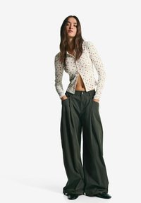 Žena s dlouhými tmavými vlasy má na sobě button-up krémový top s růžovým květinovým vzorem a široké tmavězelené kalhoty, má ruce v kapsách.