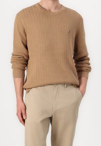 Pull en maille texturée beige avec un col rond, des poignets et un ourlet côtelés. Petit logo sur la poitrine. Porté avec un pantalon de couleur claire.