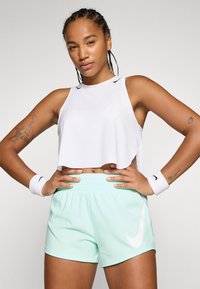 Bijela skraćena sportska majica bez rukava s crnim Nike logotipom; mint zelene kratke hlače s bijelim Nike logotipom; bijeli zavoji za zapešća. Glatka tekstura, sportski dizajn.