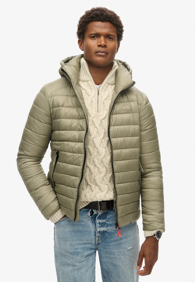 Giacca puffer verde oliva con trapuntatura orizzontale, cappuccio aderente e due tasche con zip. Indossata sopra un maglione a coste color crema e jeans.