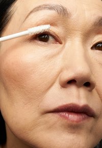Scopino per mascara con manico bianco tenuto vicino a un occhio, che mette in evidenza ciglia arricciate e una texture della pelle liscia, con un focus su tonalità naturali.