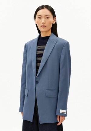 Femme aux cheveux noirs raides porte un blazer bleu clair sur un pull rayé bleu marine, sur un fond blanc uni.