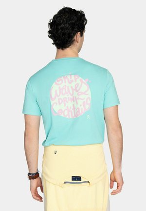 Hombre con cabello rizado de espaldas, llevando una camisa turquesa con texto rosa "Skip Waves Drink Cocktails" dentro de un círculo. Prenda de color amarillo pálido anudada a la cintura.