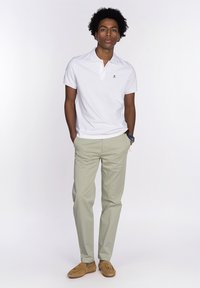 Camisa polo blanca de manga corta, pantalones chinos color caqui y mocasines marrones. El modelo está de pie con las manos en los bolsillos, el cabello arreglado de forma natural.