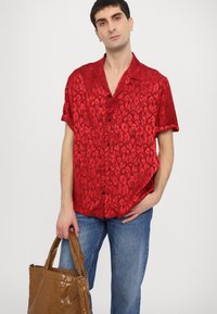 Rode T-shirt met korte mouwen, voorzien van een luipaardprint, een gladde textuur en zwarte knopen; gecombineerd met blauwe jeans en een bruine handtas.