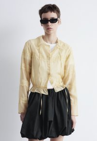 Yellow sheer blouse with lace details, button closure, and drawstring waist. Paired with a black voluminous skirt. Sunglasses in contrast.