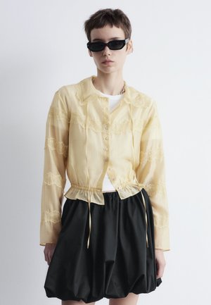 Gelbe durchsichtige Bluse mit Spitze, Knopfverschluss und Kordelzug in der Taille. Kombiniert mit einem schwarzen voluminösen Rock. Sonnenbrille in Kontrast.