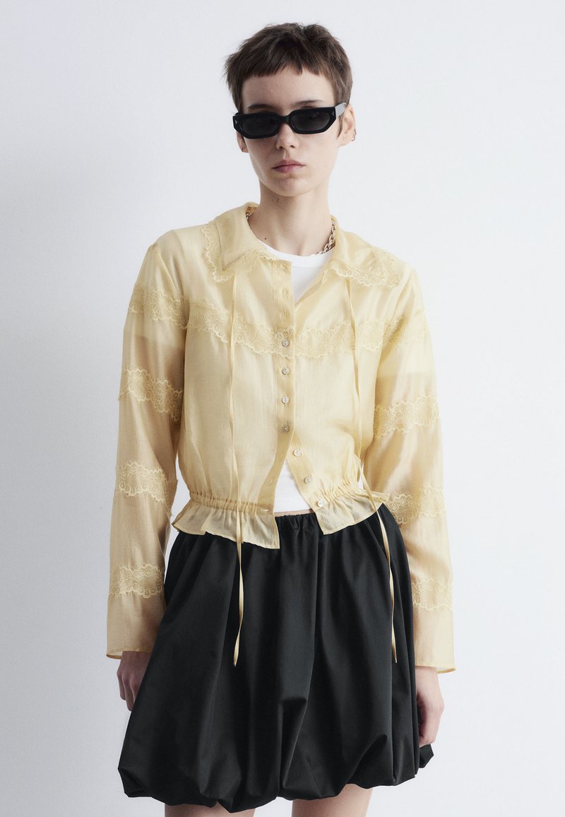 Yellow sheer blouse with lace details, button closure, and drawstring waist. Paired with a black voluminous skirt. Sunglasses in contrast.