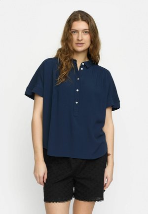 Marineblaues Kurzarmblusenshirt mit Knopfverschluss und Raffung, kombiniert mit schwarzen Shorts aus Lochstickerei mit gewelltem Saum.
