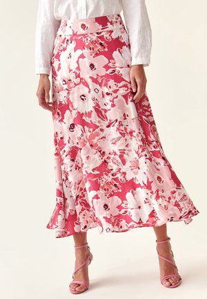 Frau trägt einen mittellangen Rock mit rosa-weißen Blumenmuster, eine weiße Bluse und rosa Riemchen-Sandaletten mit hohen Absätzen, steht vor einem einfarbigen Hintergrund.