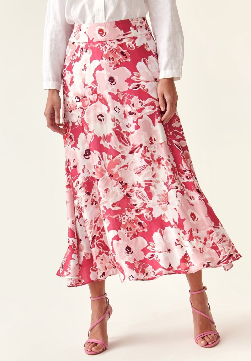 Femme portant une jupe midi fleurie rose et blanche avec un chemisier blanc et des sandales à talons hauts roses à brides, debout devant un fond uni.