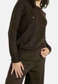 Femme portant un pull en maille pailleté marron foncé et un pantalon marron assorti, une main posée sur la hanche et l'autre sur la cuisse.