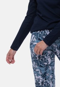 Person som bär en marinblå långärmad topp och byxor med blå blommönster, med ena handen avslappnad och den andra lätt böjd.