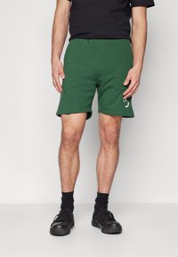 Pantaloni corti in cotone verde con fascia in vita a costine e orli laterali. Presentano un logo bianco sulla coscia sinistra. Indossati con scarpe da ginnastica nere e calze.