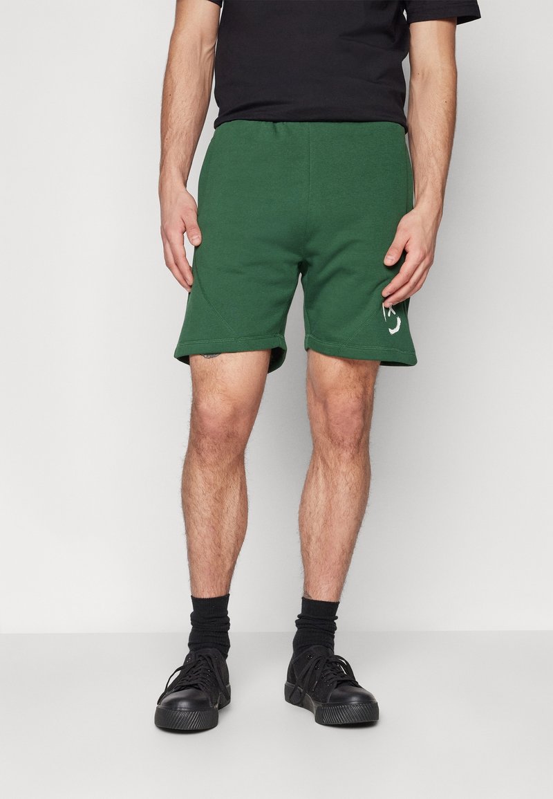 Pantaloni corti in cotone verde con fascia in vita a costine e orli laterali. Presentano un logo bianco sulla coscia sinistra. Indossati con scarpe da ginnastica nere e calze.