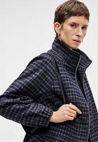 Ναυτικό και γκρι καρό σακάκι με ψηλό λαιμό, μπροστινό φερμουάρ και ελαστικούς μανδύες; συνδυασμένο με μια μαύρη τσάντα που κρατά το μοντέλο.