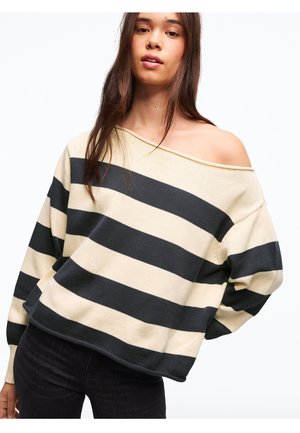 Junge Frau trägt einen schulterfreien, locker sitzenden Pullover mit cremefarbenen und schwarzen horizontalen Streifen sowie schwarze Hosen, mit langem, glattem braunem Haar.