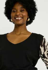 Top de punto negro con escote en V y mangas cortas que presenta un patrón abstracto de print animal en crema y marrón. Textura suave, diseño ajustado.