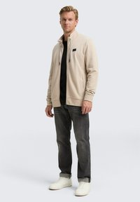 Giacca zip-up beige chiaro con colletto alto, texture a coste, tasche laterali, abbinata a una maglietta nera, jeans grigi e sneakers bianche.