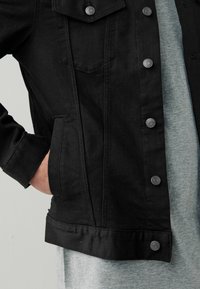 Chaqueta de mezclilla negra con cierres de botón, bolsillos frontales y un acabado texturizado. Llevada sobre una camisa gris, mostrando un look en capas.