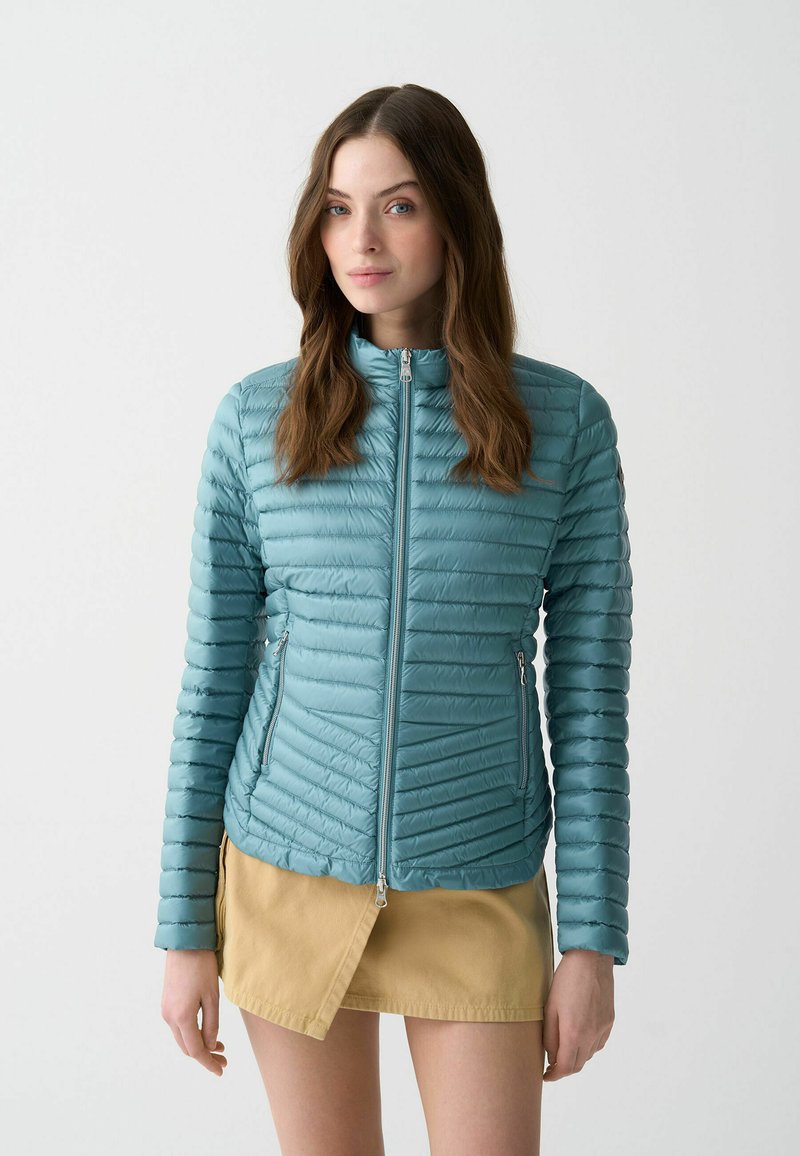 Hellblaue Steppjacke mit horizontaler Steppung, Frontreißverschluss und seitlichen Taschen. Kombiniert mit einem khakifarbenen Rock posiert das Modell vor einem weißen Hintergrund.