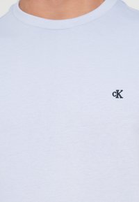 Pogled od blizu na osebo, ki nosi svetlo modro bombažno majico s majhnim črnim vezenim logotipom "cK" blizu prsnega dela.