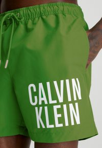Grønne svømmeshorts med en glat tekstur, med en hvid "CALVIN KLEIN" logo. Elastisk talje med snøre og sidelommer.