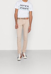 Beige slim-fit-housut, valkoinen graafinen t-paita, jossa on "JACK & JONES" -teksti, ruskea punottu vyö ja moniväriset lenkkarit, joissa on valkoiset pohjat.