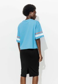 Femme aux cheveux courts et bouclés portant une chemise bleue à manches courtes avec des rayures et une jupe noire au genou avec une fente à l'arrière, se tenant de profil.