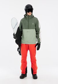 Snowboarder iført en grøn jakke med grå accenter og røde bukser, holder et sort snowboard med et hvidt brandlogo, og har handsker og en hjelm på.
