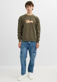Sudadera verde oliva con un logo en el frente, combinada con vaqueros cargo azules con bolsillos laterales y zapatillas blancas.