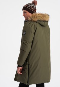 Parka verde oliva con capucha de piel sintética, que presenta un cierre de cremallera, puños acanalados y un parche con logo en el hombro.