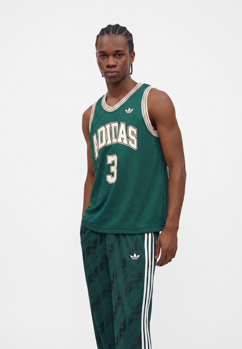 Ung mand med fletninger iført mørkegrøn Adidas basketballtrøje og matchende træningsbukser, stående mod en simpel hvid baggrund.