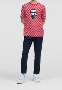 Camiseta roja de manga corta con un patrón repetido y un personaje de dibujos animados en blanco y negro, combinada con pantalones oscuros azules y zapatillas blancas.