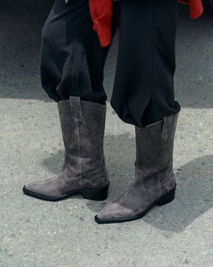 Persona que lleva botas de ante gris hasta media pantorrilla con tacones bajos y pantalones negros sueltos, de pie sobre pavimento gris.