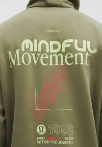 Hoodie vert olive avec "MINDFUL Movement" en texte vert clair et lignes ondulées rouges. Comprend le logo "lululemon" et l'année "1998" en bas.