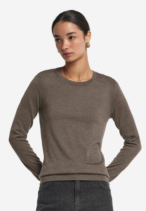 Frau mit zurückgebundenem dunklem Haar, trägt goldene Creolen, einen braunen Pullover mit langen Ärmeln und dunkle Jeans, steht mit den Händen hinter dem Rücken.