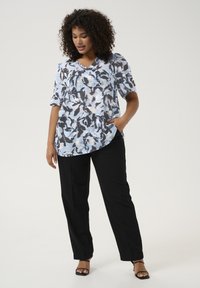 Blouse fluide à motif dans des tons de bleu clair, gris et noir ; décolleté en V avec des manches courtes et un ourlet arrondi. Associée à un pantalon noir.