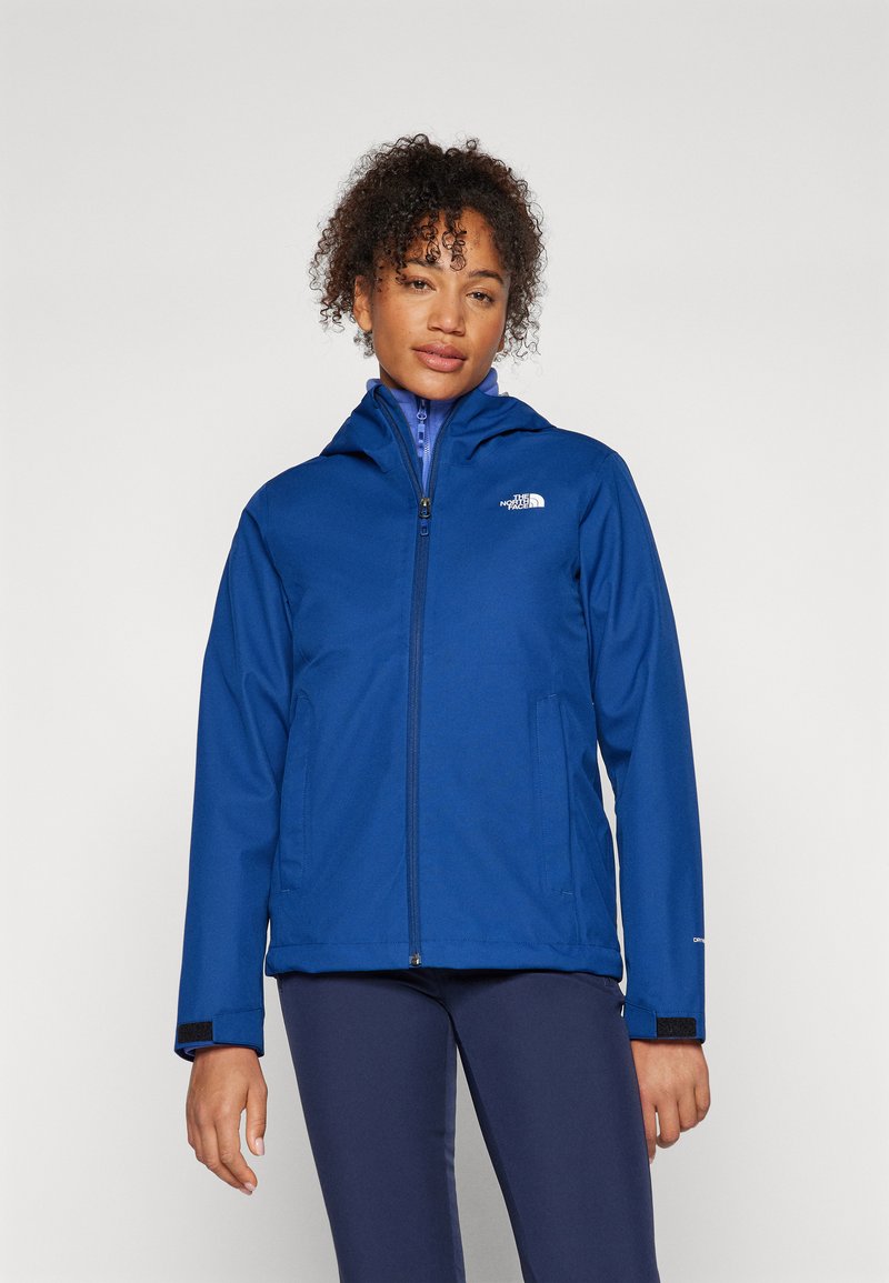 Modra softshell jakna s kapuco, zadrgo in dvema stranskima žepoma; ima logotip na prsih in Velcro zapestja za prilagajanje.