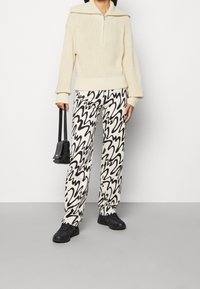Pullover in maglia crema con zip a metà, e pantaloni larghi a motivi bianco e nero. Gli accessori includono una piccola borsa nera e scarpe nere.
