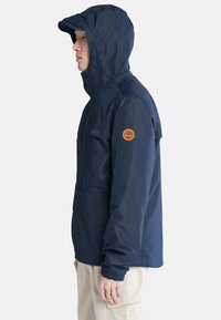 Giacca impermeabile blu navy con cappuccio, polsini elastici e un grande patch con logo sulla manica. Tessuto leggero, design pratico.