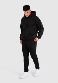 Felpa nera con zip dal tessuto morbido, abbinata a pantaloni jogger neri coordinati. Entrambi hanno una vestibilit�à comoda e sono indossati con sneakers bianche.