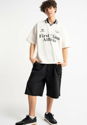 Młoda osoba z kręconymi włosami nosząca białą, oversize'ową koszulę z napisem „First Class Athlete”, czarne długie spodenki oraz czarne tenisówki z białymi skarpetkami.