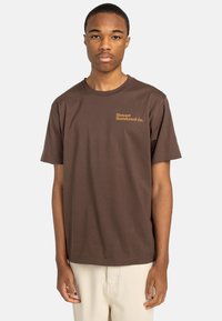 Camiseta de algodón marrón de mangas cortas y cuello redondo. Presenta una impresión de logotipo en dorado en el pecho izquierdo y un diseño limpio.